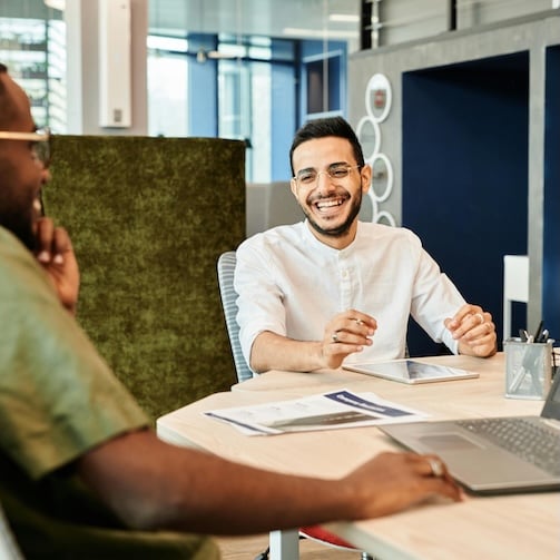Two people sitting at a table in a modern office, discussing documents and working with a laptop and tablet.