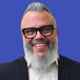Professional headshot of a Fabian Campos wearing eyeglasses, a dark blazer, and a light shirt, photographed against a blue background in a close-up portrait composition.