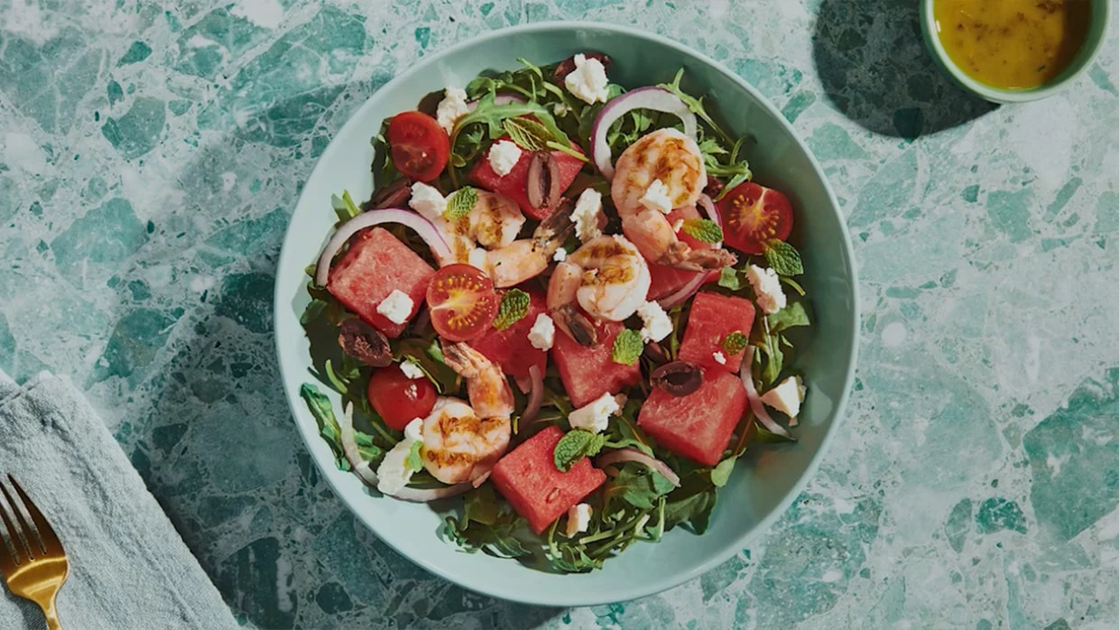 A bowl of salad with watermelon, shrimp, cherry tomatoes, feta, olives, red onion, mint, and arugula on a marble table.