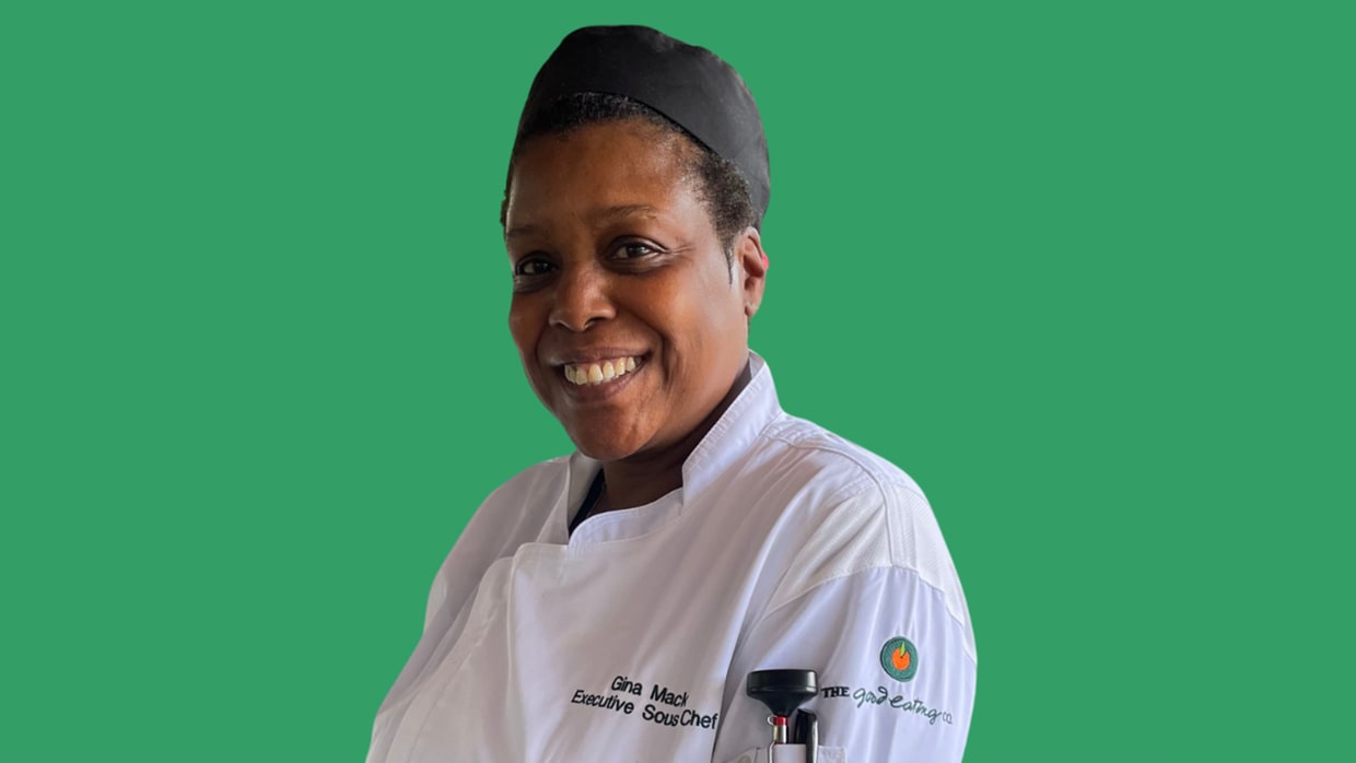 Chef Gina Mack wearing a white chef's coat with "Executive Sous Chef" embroidered on it, standing against a green background.