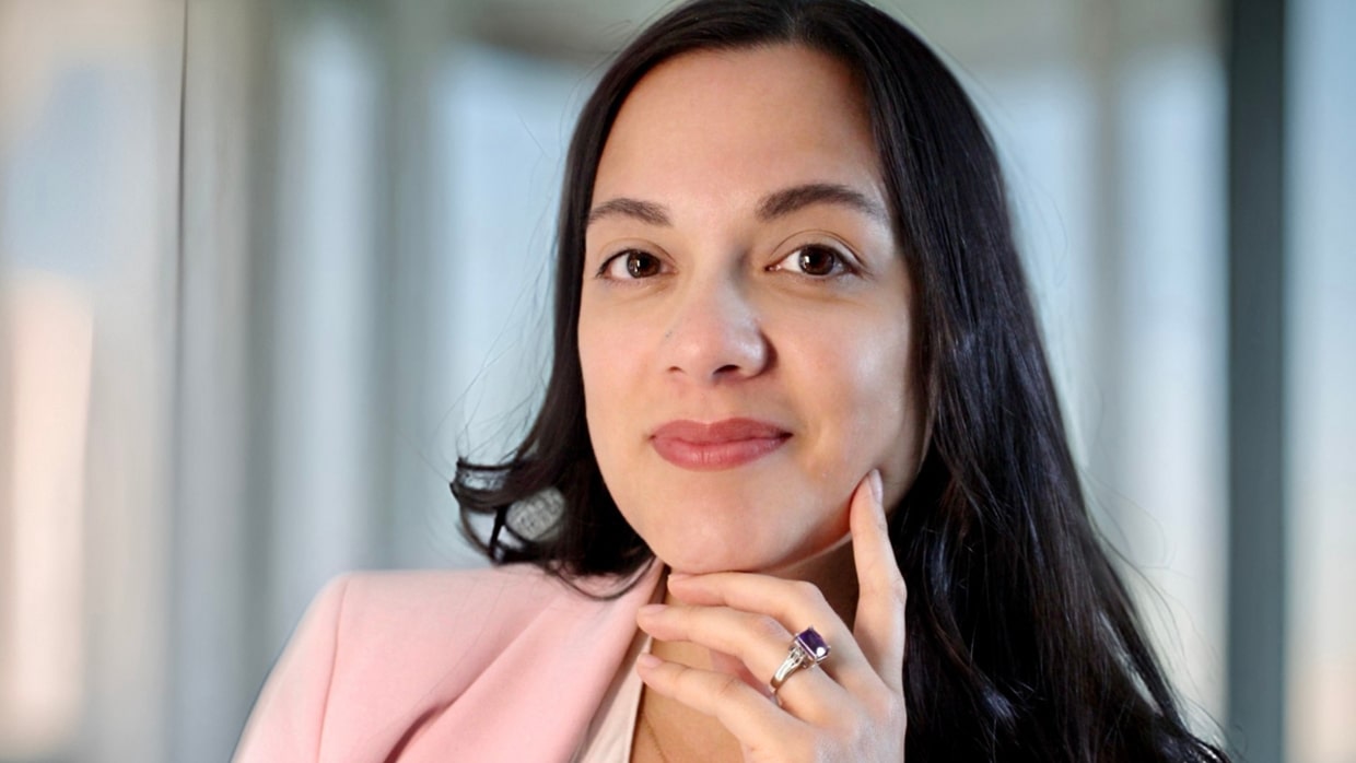 Person with long dark hair in light pink blazer, wearing a purple gemstone ring