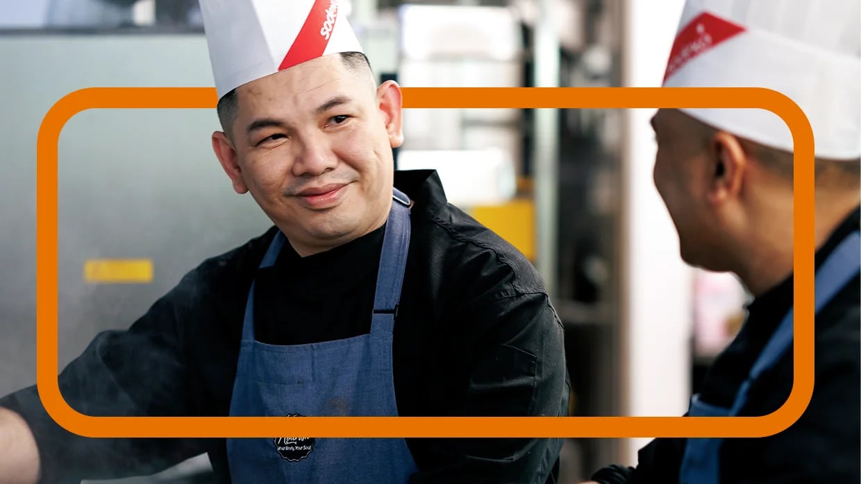 Two chefs in Sodexo hats and aprons smile while working together in a kitchen setting.