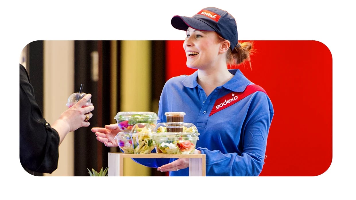 Smiling Sodexo worker hands over packaged salads and drinks to customer in bright, modern setting.