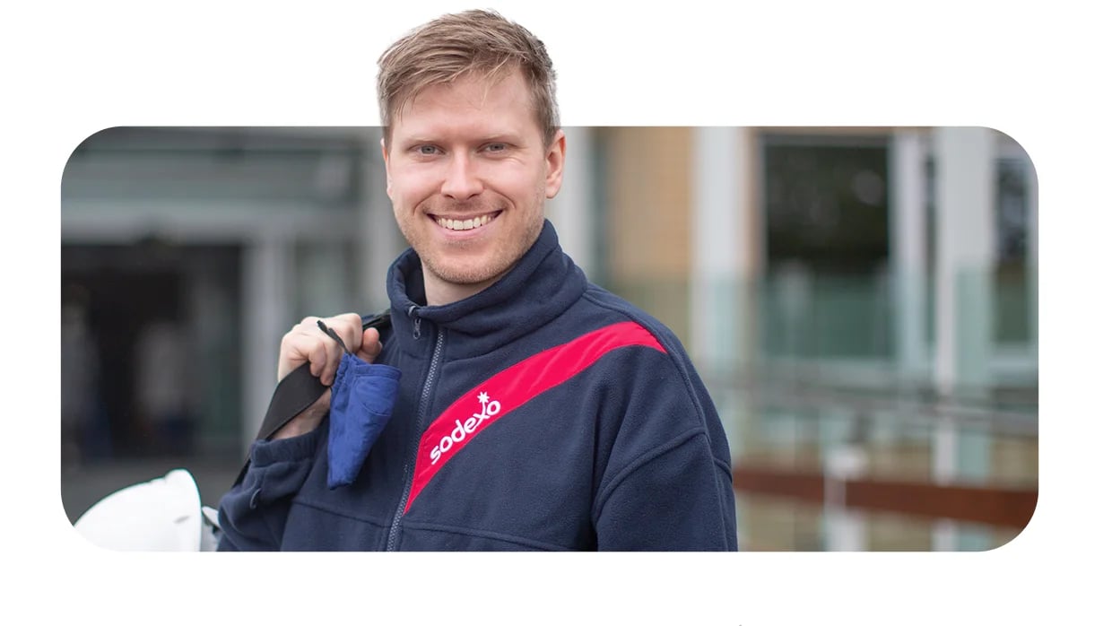 Sodexo worker holds blue cloth and white helmet, wearing branded jacket with red stripe in work setting.