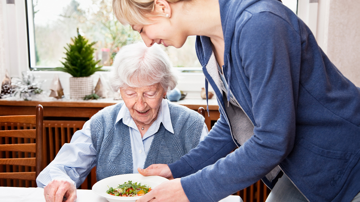 A senior woman sitting at a table, being served a meal by a young caregiver