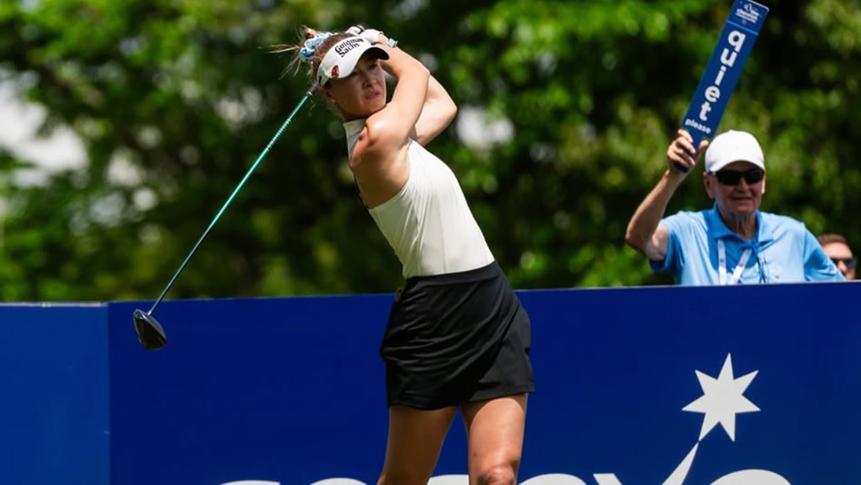 Golfer follows through on a tee shot while an official stands nearby holding a quiet sign.