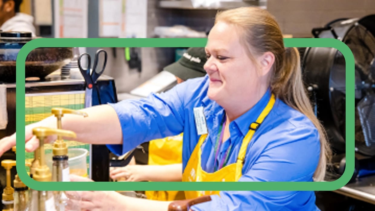Smiling food service worker in yellow apron pumps sauce into cup at busy prep station.