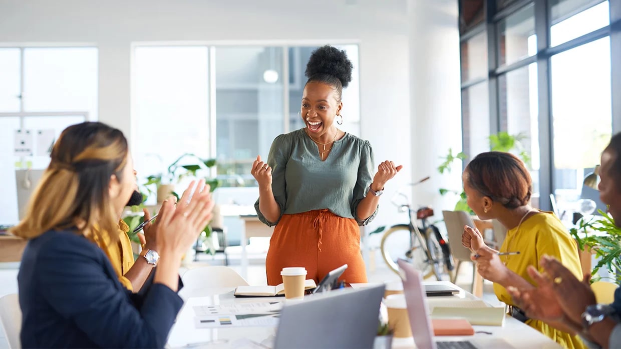 A person stands and speaks while others sit and applaud in a modern office, suggesting a presentation or recognition moment.