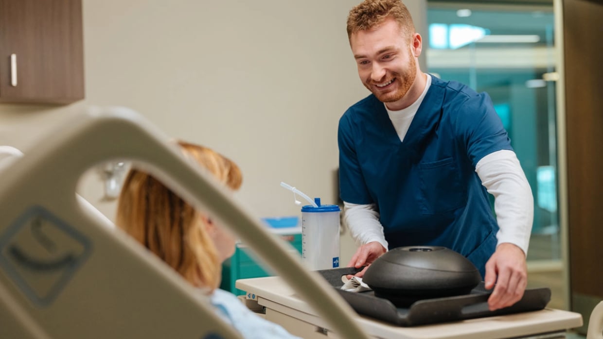 A nurse in blue scrubs brings a covered meal tray to a patient in a hospital room