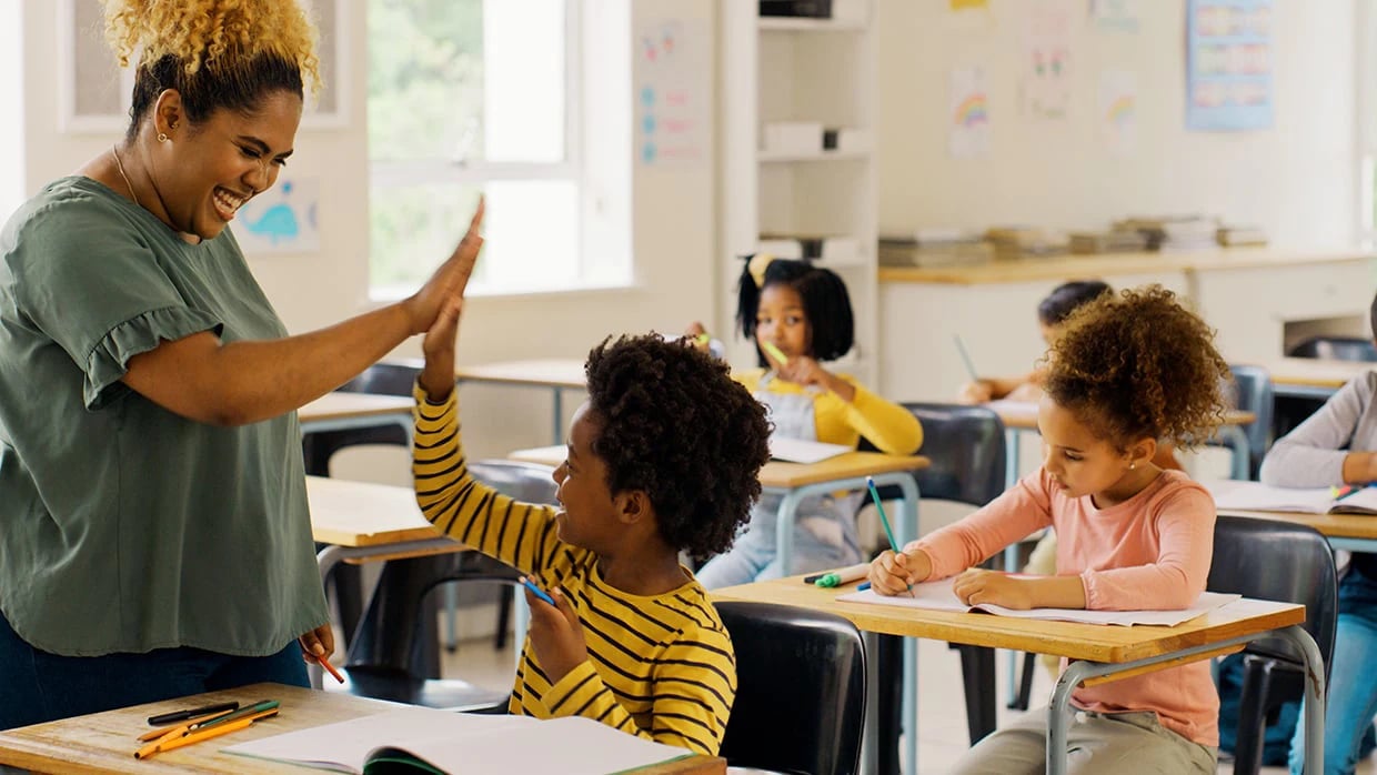 Teacher high-fiving a smiling student in a bright classroom as other students work at desks.