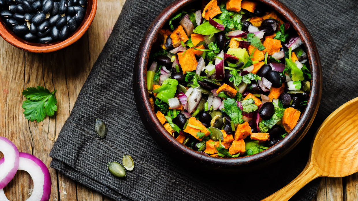 A colorful salad with black beans, sweet potatoes, red onions, green peppers, cilantro, and pumpkin seeds, displayed on a dark cloth napkin over a rustic wooden table.