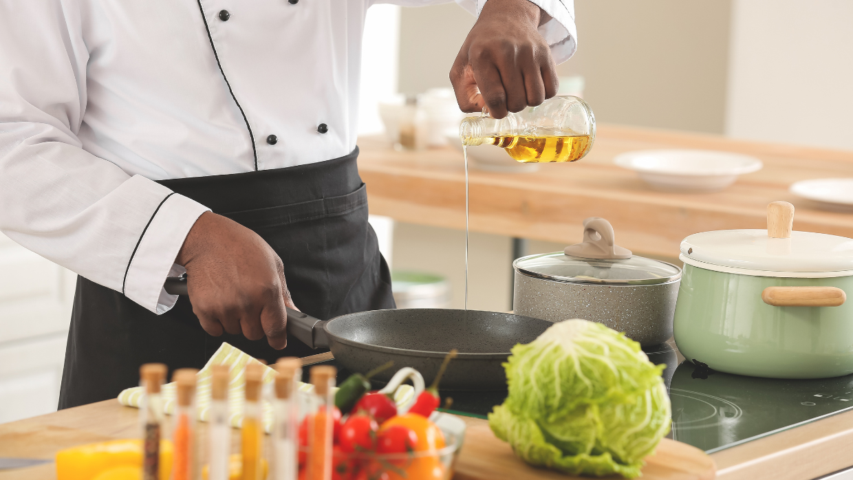 Image of a chef in a white jacket and black apron pouring oil into a frying pan in a busy kitchen with fresh ingredients. 