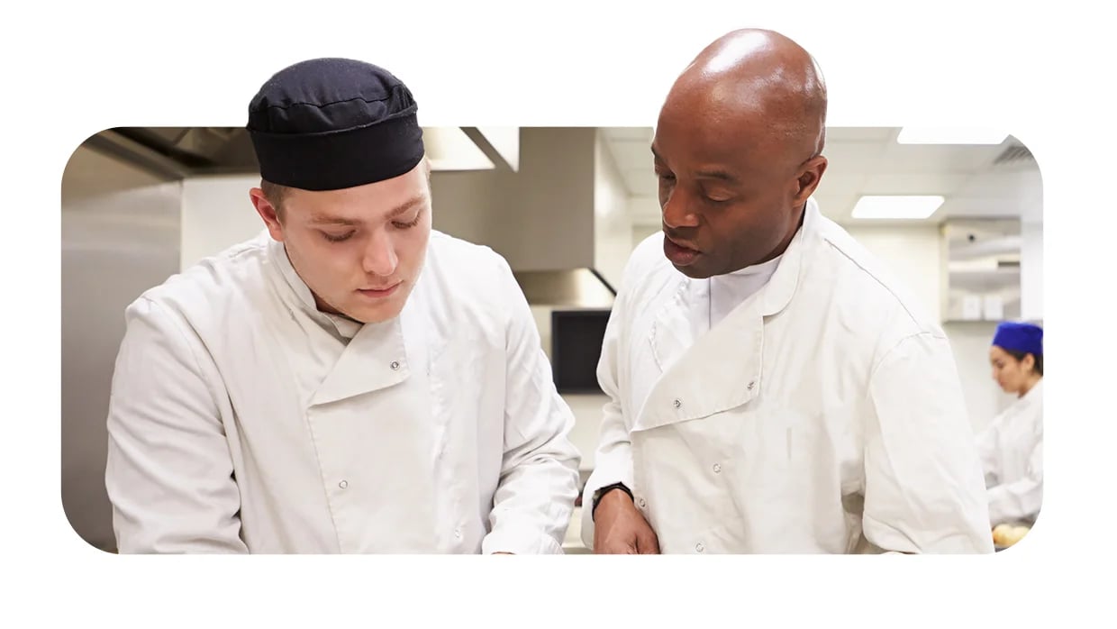 Chef instructs young cook in white uniform during food prep in professional kitchen.