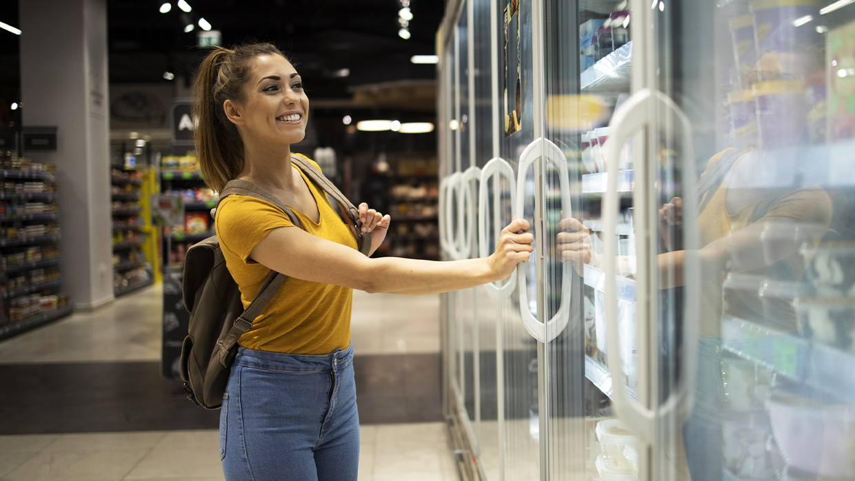 A female student, wearing a backpack, smiles while browsing the refrigerated section of a convenience store.