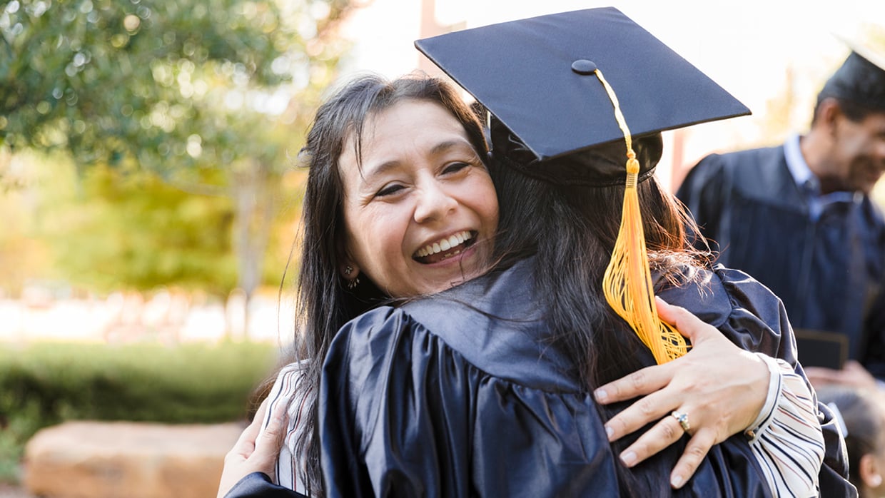 Student wearing a graduation cap and gown, hugging her smiling mom.