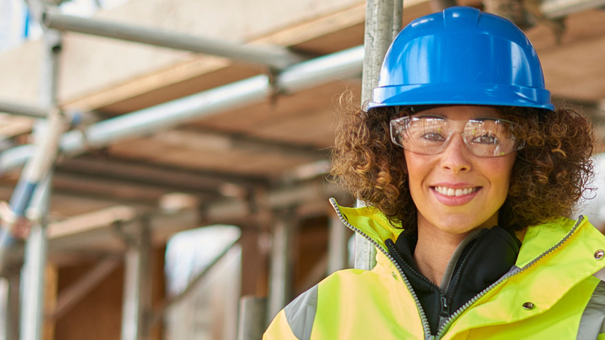 A woman wearing a safety vest and hard hat, standing confidently at a construction site.