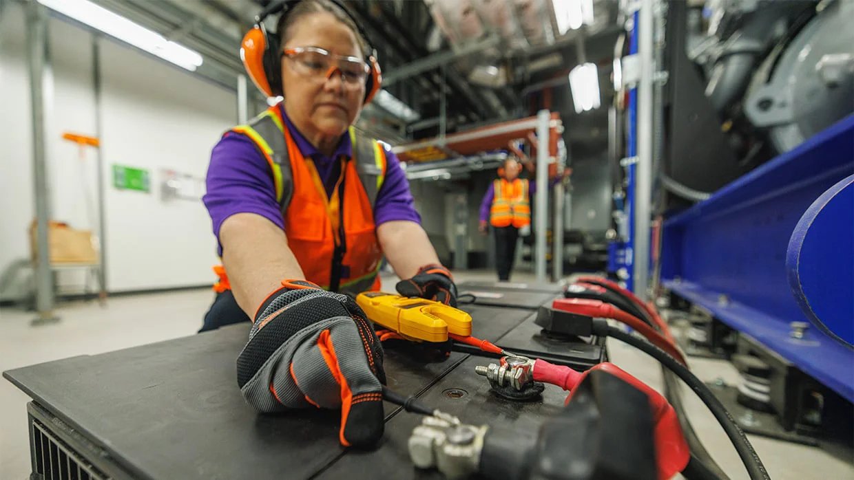 Worker in safety gear tests a large battery with a multimeter in an industrial environment.