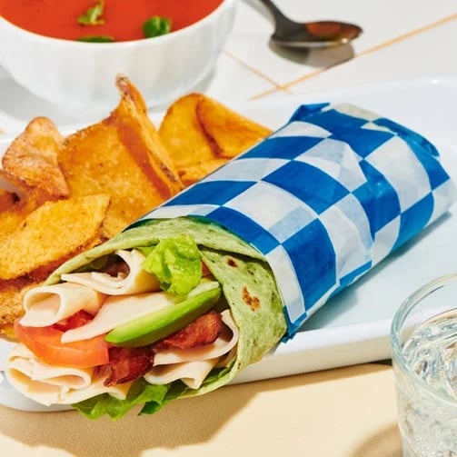 Turkey, lettuce, tomato and avocado wrap in blue-checkered paper, with potato wedges and tomato soup.