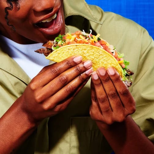 Hand holding taco filled with seasoned meat, lettuce, cheese, and tomatoes, ready to take a bite.