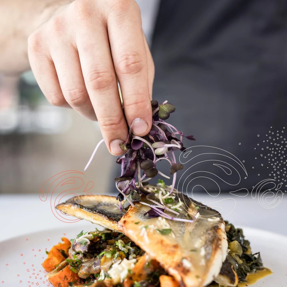 A hand garnishes a plated fish dish with microgreens, colorful veggies, and sauce swirls on a white plate.