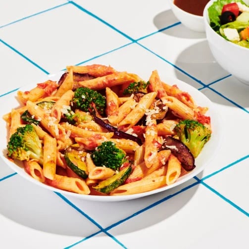 Penne pasta with broccoli, zucchini, eggplant, tomato sauce, and cheese beside salad on white tiled surface.