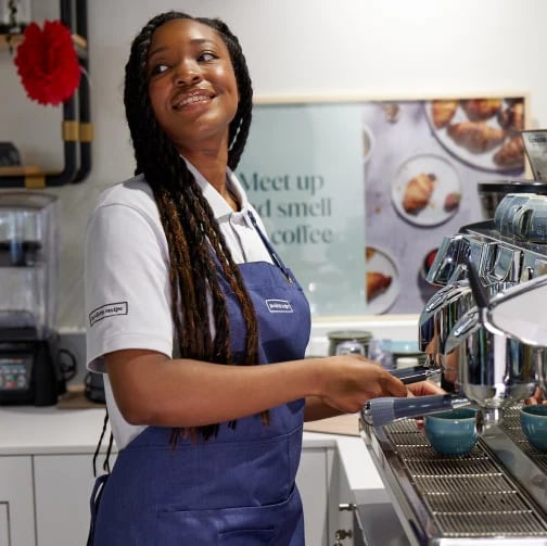 Barista in blue apron uses espresso machine in cafe, with blender and “Meet up and smell the coffee” poster behind.