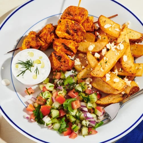 Plate with chicken skewers, seasoned potato wedges topped with feta, tzatziki bowl, and a fresh veggie salad.