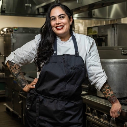 Chef in white jacket and black apron stands in a professional kitchen with visible arm tattoos, surrounded by industrial cooking equipment and large pots.