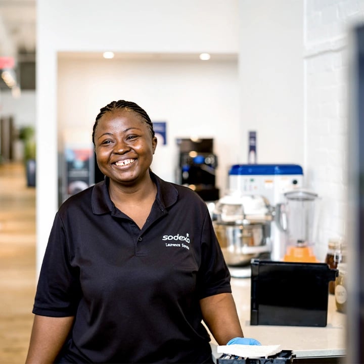 A smiling woman in a black shirt stands in front of a counter, exuding a friendly and welcoming demeanor.