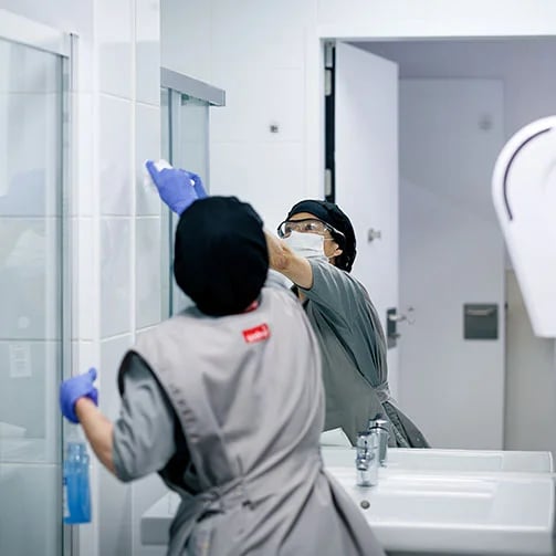 person in protective gear clean a bathroom; one wipes a mirror, the other holds a spray bottle. Provide your feedback on BizChat