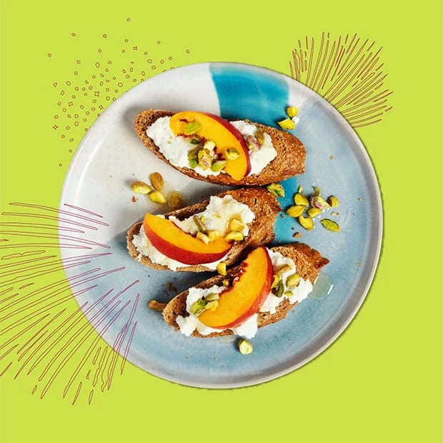 Three slices of bread topped with ricotta, peach slices, and pistachios on a blue-and-white plate.
