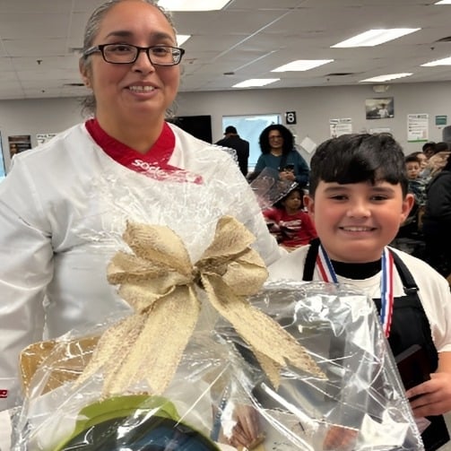 Adult and child at school event holding a wrapped gift basket, with onlookers in a classroom setting.
