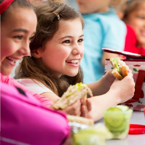 Children enjoying lunch together in a school cafeteria, seated at tables with trays of food and chatting happily.