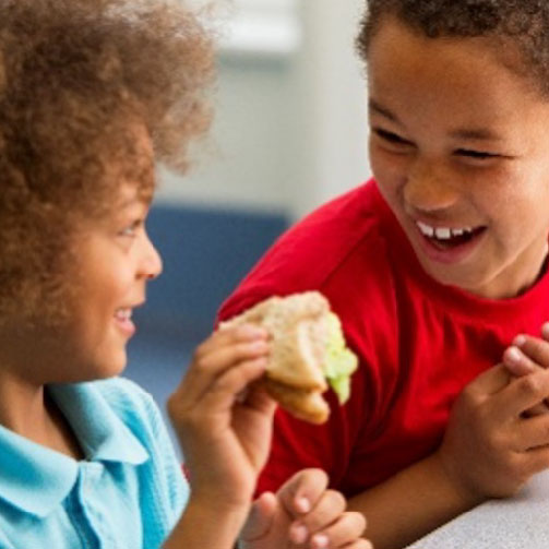 Children enjoying lunch together in a school cafeteria, seated at tables with trays of food and chatting happily.