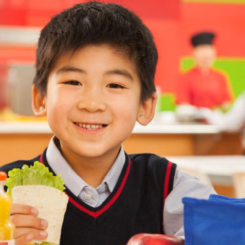 A young boy is smiling while holding a sandwich in one hand and a drink in the other.