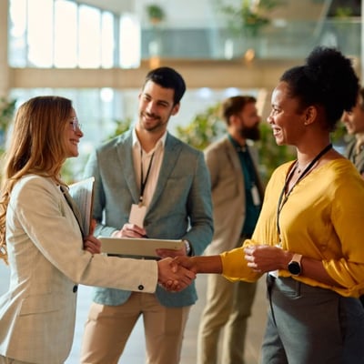Two businesswomen shaking hands at a conference, with other professionals conversing in the background.