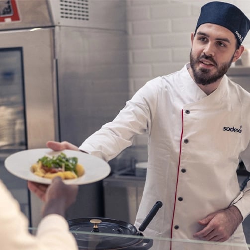A man in a white chef's coat hands a plate to a woman, both smiling in a bright kitchen setting.