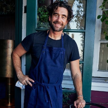 Chef Gabe Kennedy stands smiling in a doorway, wearing a dark t-shirt and a blue apron.