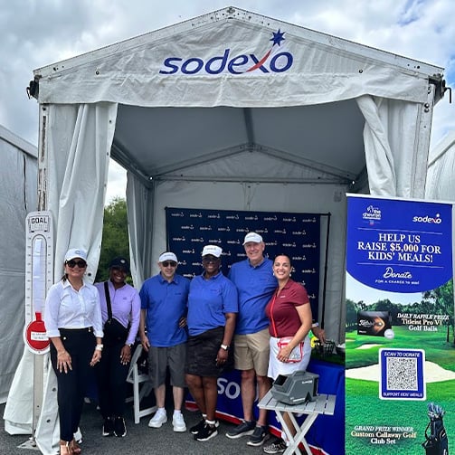 Group poses in a Sodexo event tent beside a fundraiser display supporting meals for children.