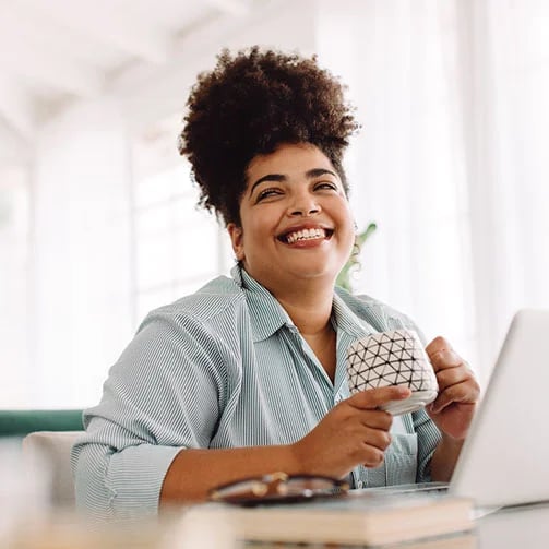 A person in a striped shirt holds a geometric-patterned mug while working on a laptop in a bright, modern workspace.