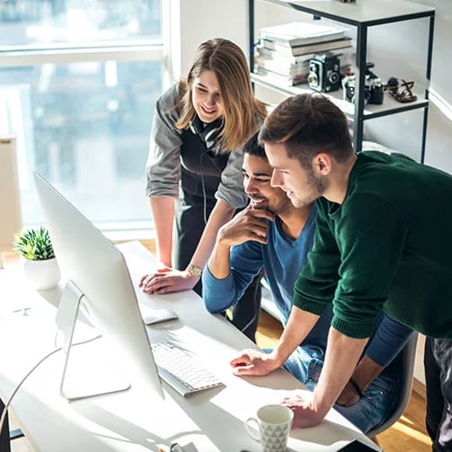 Three people collaborate at a desk, focused on a computer screen, in a brightly lit office environment.