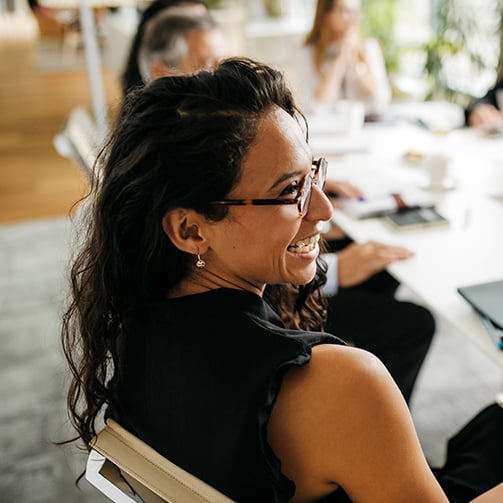 Smiling woman turns toward camera during a team meeting, with colleagues and laptops blurred in background.