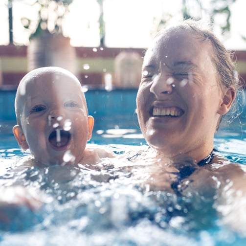 Adult holding a baby in a pool as water splashes around them. 