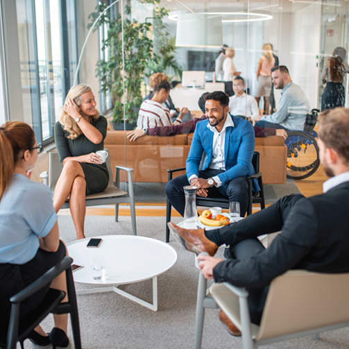 People seated in a modern office lounge having a small group discussion around a coffee table, with drinks and fruit, while others work and talk in the background.