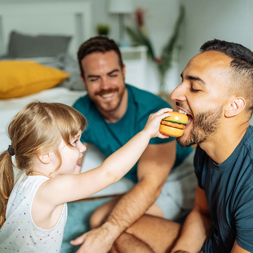 Toddler playfully offering a toy hamburger to an adult as another adult watches and smiles nearby.