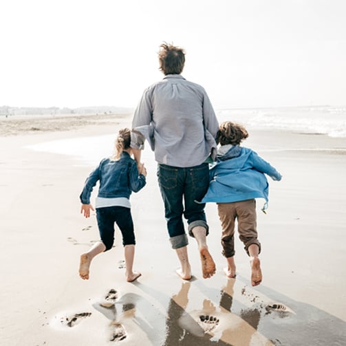 Adult and two children holding hands running barefoot along a sandy beach near the shoreline.