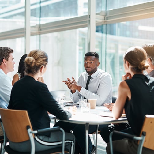 Group of colleagues sit around a table in a glass-walled office discussing during a workplace meeting.