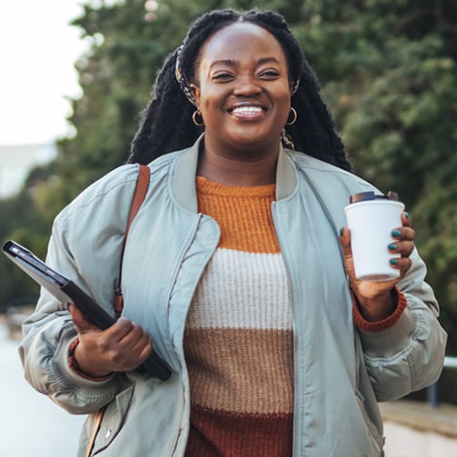Person smiling and walking outdoors on a tree‑lined path, holding a coffee cup and a tablet, wearing a striped sweater and light jacket. 