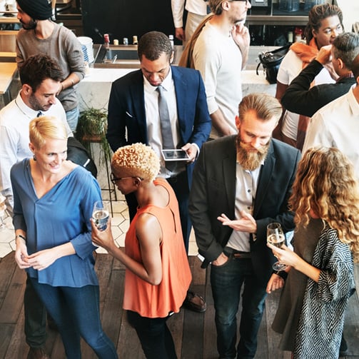 Overhead view of professionals mingling at a workplace event, standing in small groups, holding drinks, and talking in a modern indoor space with natural light.
