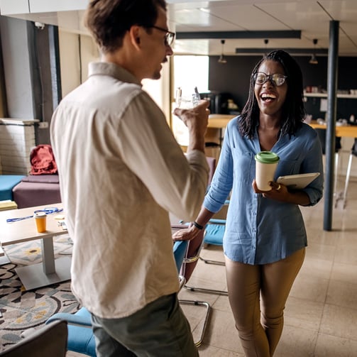 Two coworkers laughing and chatting in a modern office lounge, one holding a coffee cup and tablet.